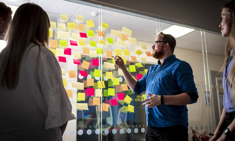 A bearded man points at a think board of post its stuck to a see through surface, while several students look on
