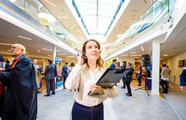 Female student standing in a busy room holding a clipboard and talking on a headset