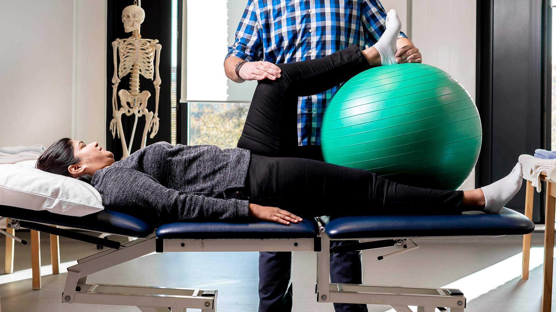 Patient lying on a physio bed with a practitioner moving patient's leg on top of a large green ball 