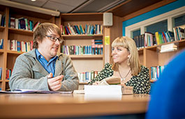 Two students sitting at a desk, talking, with bookshelves behind them