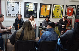 Lecturer and group of students at a boardroom style table with lots of football shirts behind them