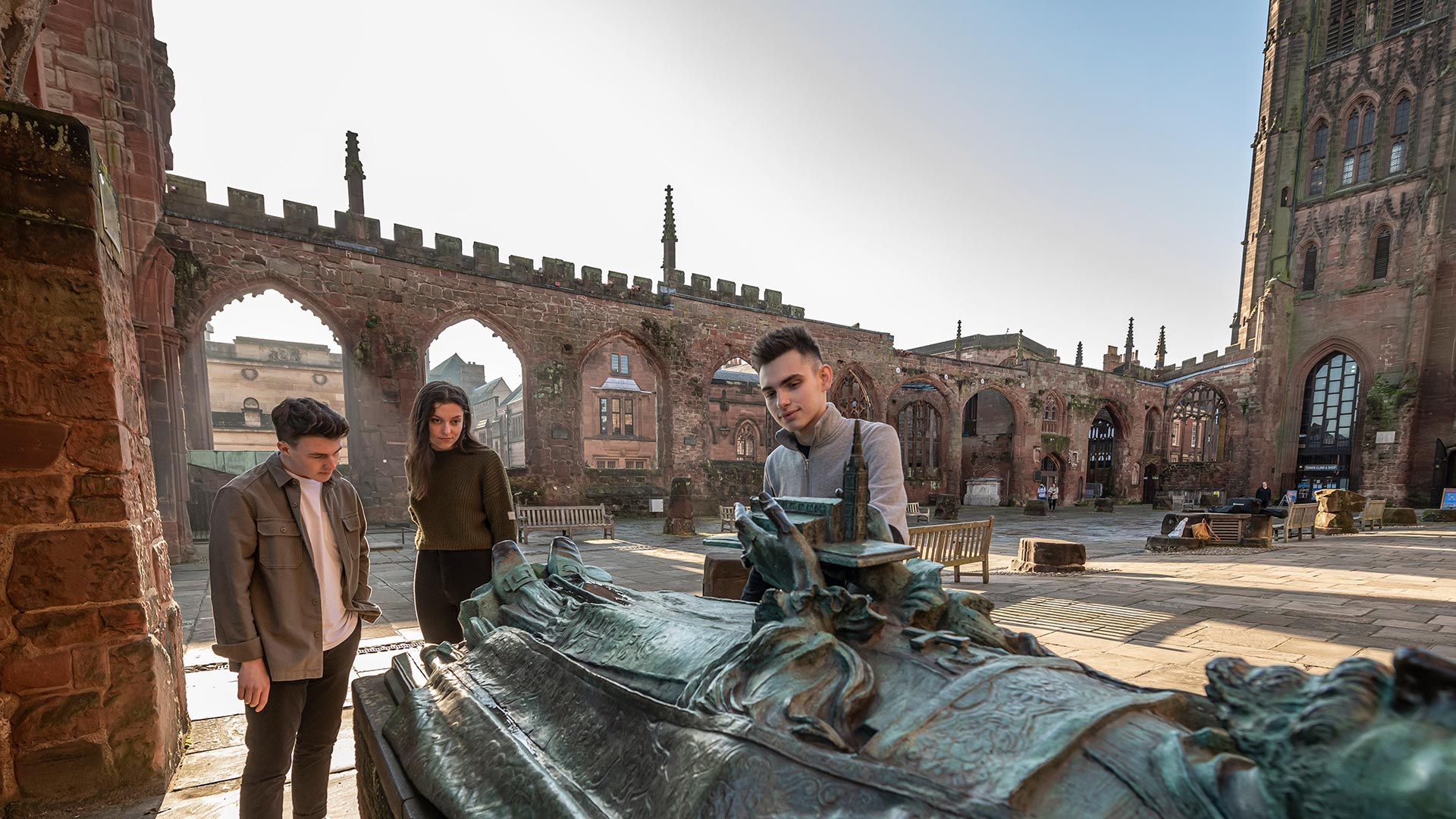 Three students in Coventry Cathedral grounds looking over a sculpture of a man laying down