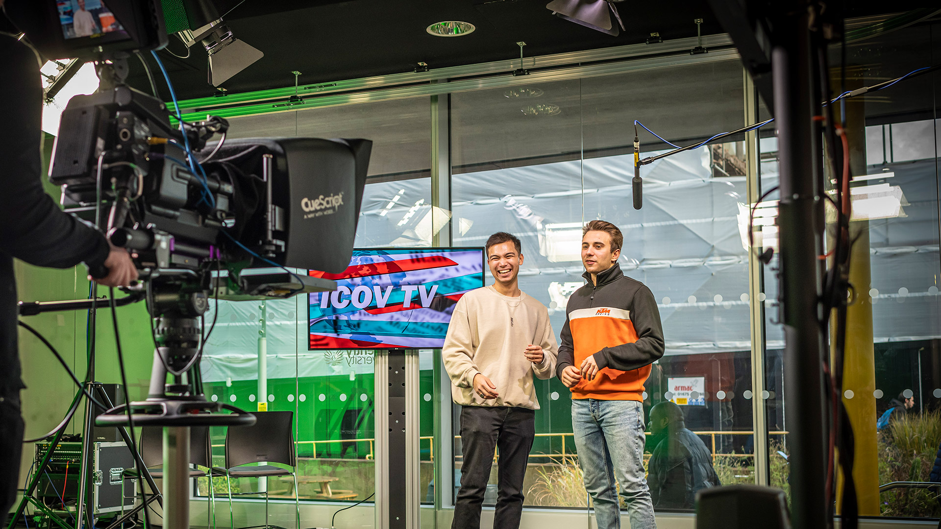 Two students standing in front of a filming camera emulating a tv studio