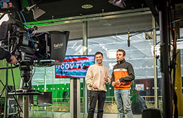 Two students standing in front of a filming camera emulating a tv studio