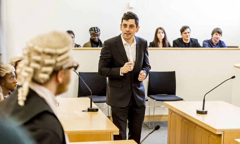 A lawyer stands in a court room, with people behind we assume make up the jury, and two judges to his left