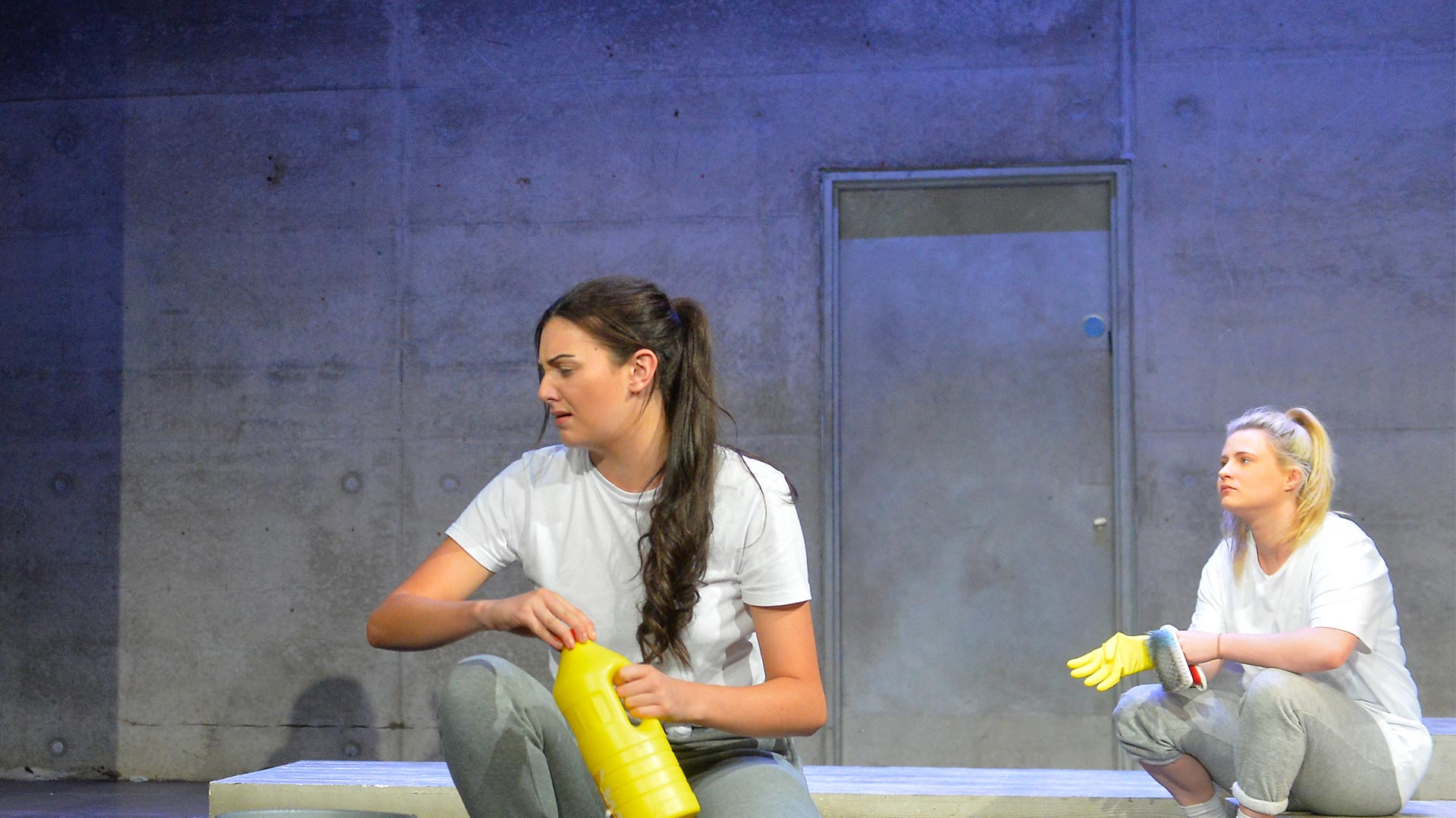 Two students acting on stage sat on the floor with a concrete wall behind and stage lighting