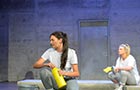 Two students acting on stage sat on the floor with a concrete wall behind and stage lighting