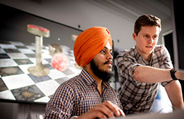 Student wearing turban looking at screen, tutor leaning over pointing at the screen