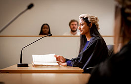 Female law student wearing robes and wig sitting in a court with notes in front of her, two people sitting behind her, blurred