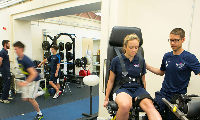 Woman using sports equipment supervised by man wearing glasses, in the background someone sprints past