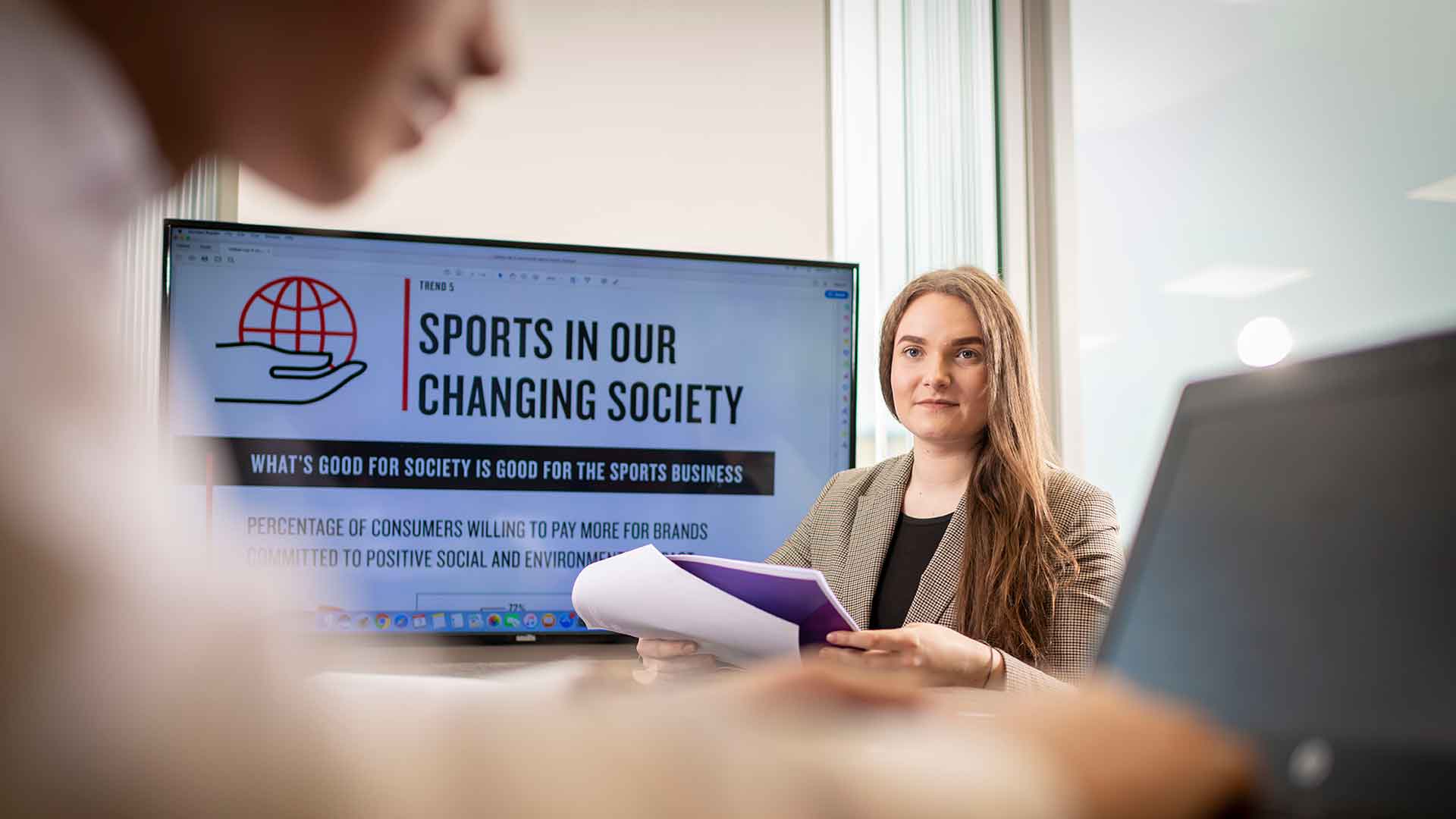 Female student sitting at a desk holding a document talking to a male student with a TV behind