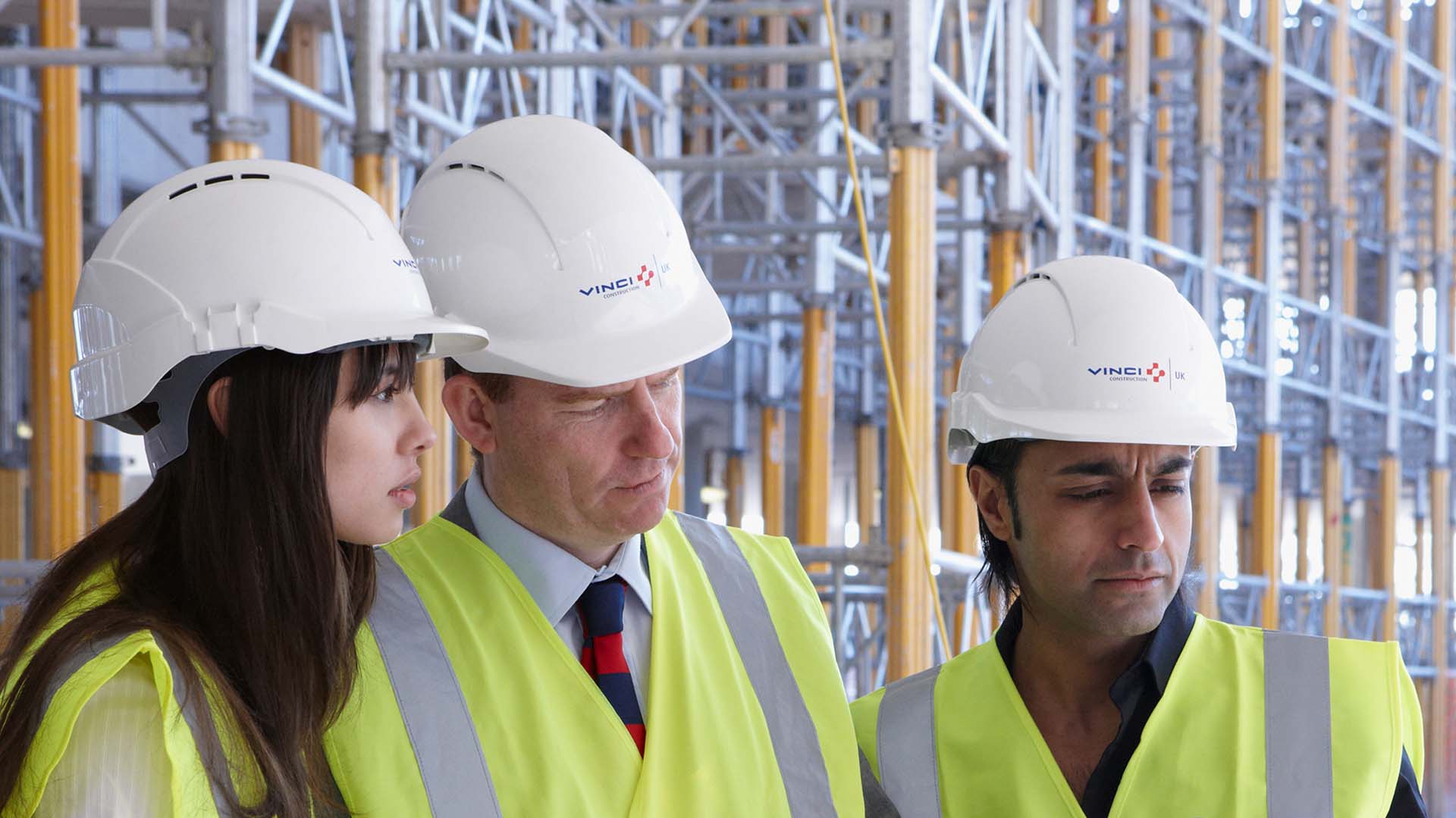 Three people with hardhats and hi-vis vests outside scaffolding
