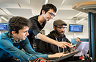 Three people lean over a screen, the middle one standing and pointing at the screen.