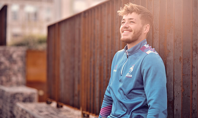 Student Dan Kiernan wearing sports kit, sitting against a wall, smiling and looking up into the sunlight