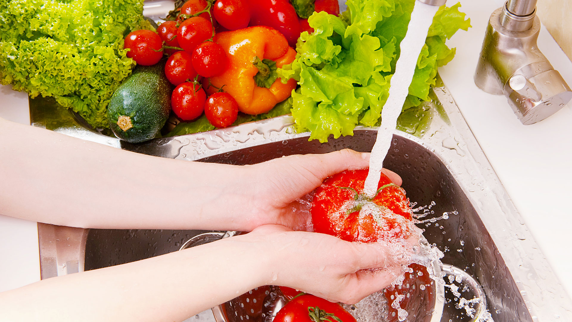 Arms and hands washing a vegetable in a sink with the tap running and other vegetables in the side