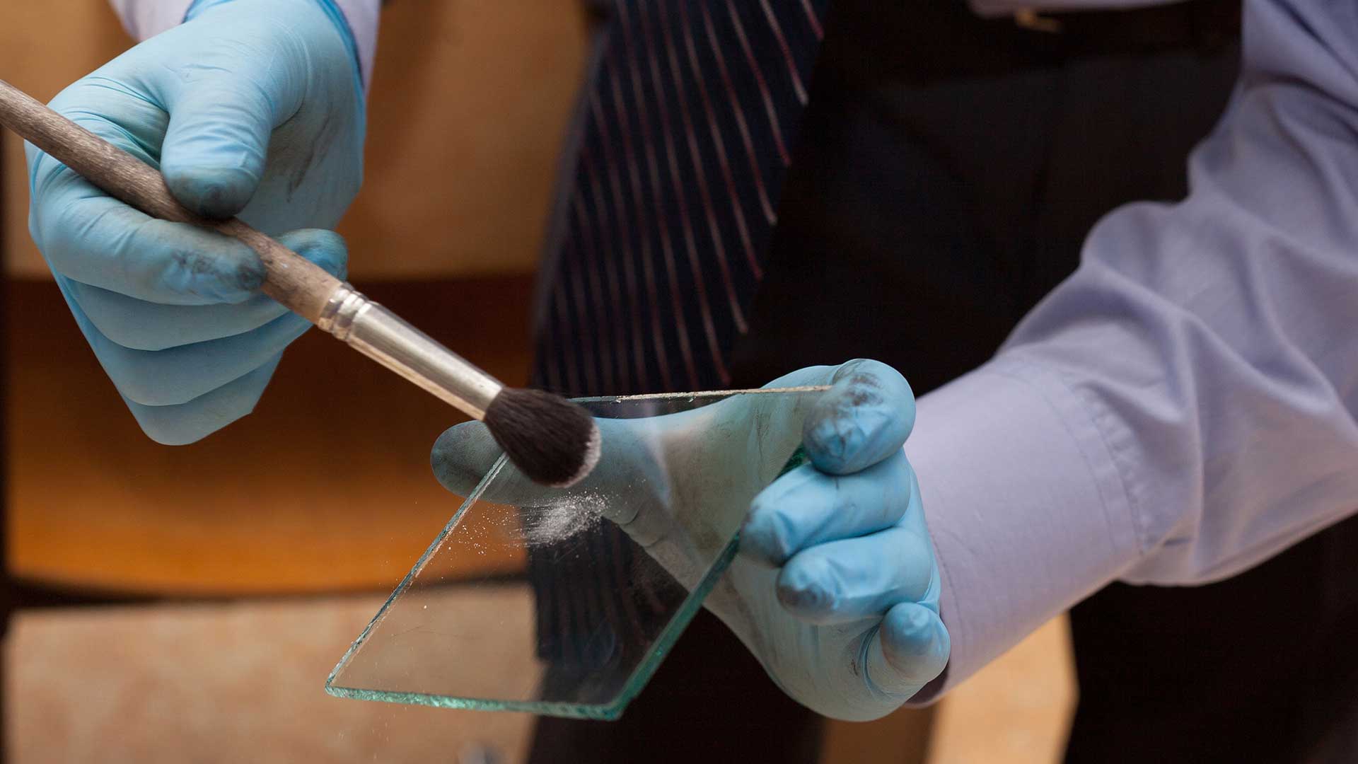 Up-close hands in blue gloves and a shirt holding a piece of glass with a brush testing carrying out an investigation