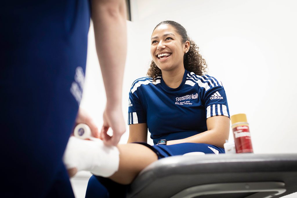 A smiling student looks up at the person just out of shot, who is wrapping her ankle in a bandage