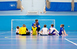 Students sat together on a sports hall blue floor with a tutor in front teaching