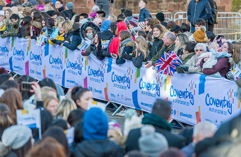 Crowds of people behind railings holding flags waiting to see the Duke and Duchess of Cambridge