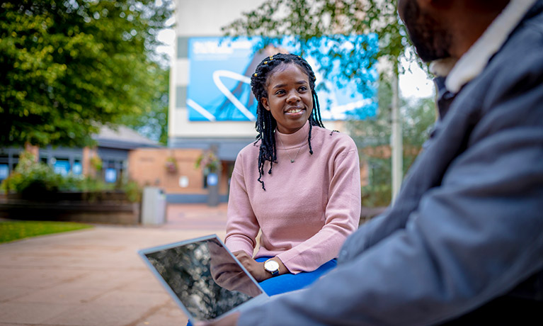 Female student sat outside talking a male student with his laptop