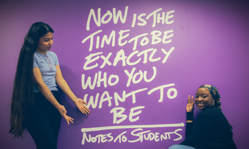 Two students standing next to large wall art that reads now is the time to exactly who you want to be
