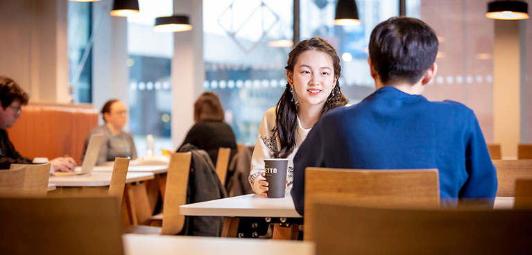 Male and female student sitting in a cafe talking with people sat in the background