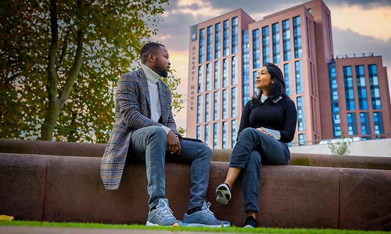male and female student sitting on a wall outside with the library in the background