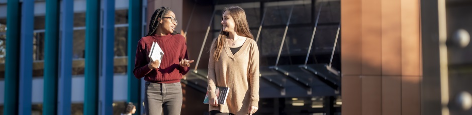 A couple of students walking outside in the sun with a university building in the background