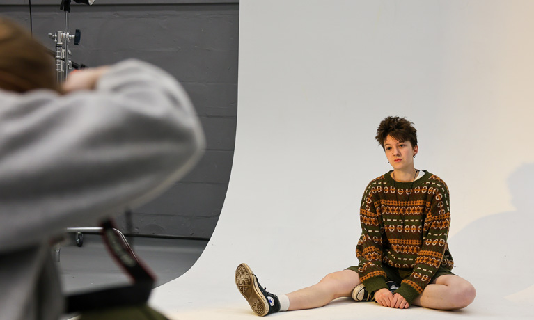 A student sitting down while another student takes her photograph against a white backdrop in the photography studio