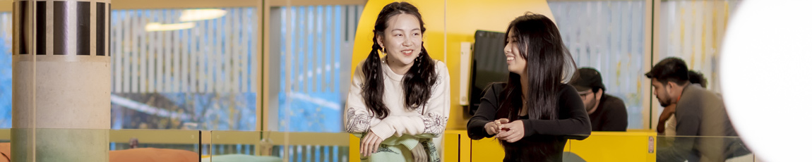 Two international students smiling and looking over the side of a glass balcony.