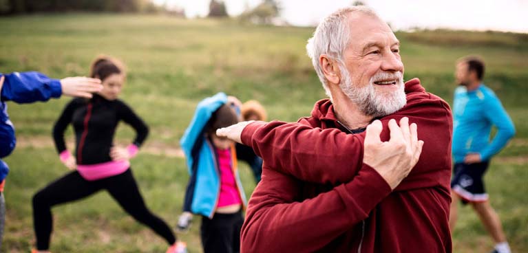 older man stretching his arm with others stretching in an open space