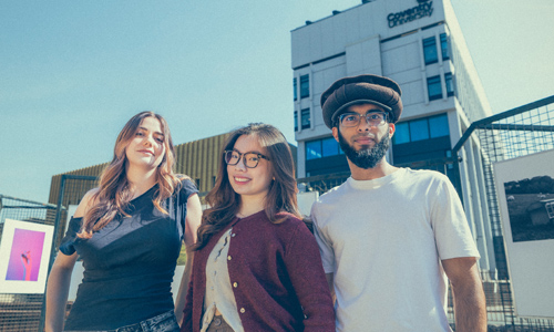 Three students stood in Coventry University's Starley Gardens on a sunny day, with two campus buildings in the background