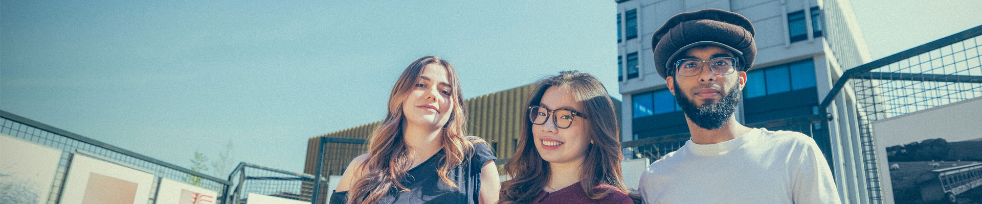 Three students stood in Coventry University's Starley Gardens on a sunny day, with two campus buildings in the background