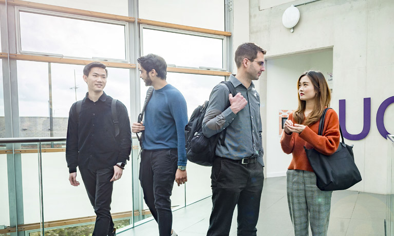 three male students and one female student standing talking at the top of the stairs inside a building