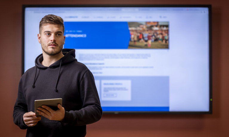 Male student holding a tablet presenting with work on a screen behind him
