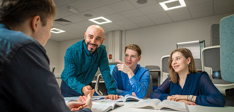 Lecturer stood at a table talking to a group of students sat around the table