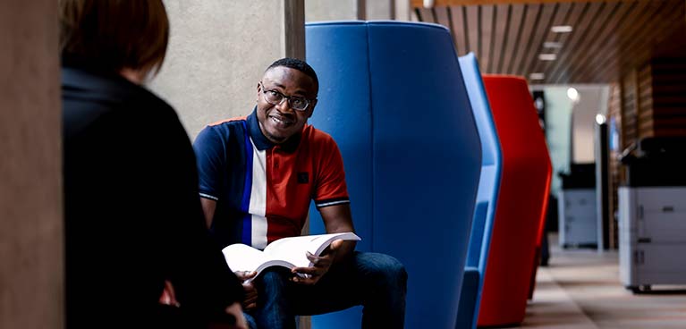 male student sitting down holding a book chatting to another person with their back to the camera