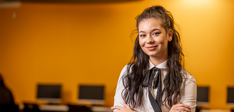 A student smiling and crossing her arms in a portrait with a bright yellow wall and computer screens behind