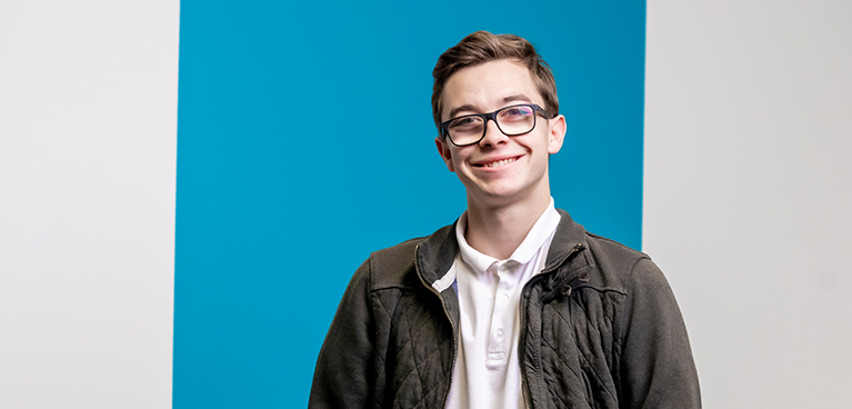 A student wearing glasses and jacket smiling at the camera in a portrait