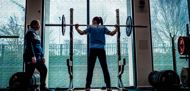 Person lifting a weight on their shoulders with instructor looking  on