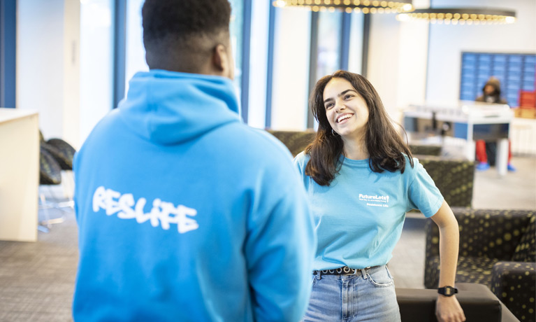 Maria Fernandez in a Halls of Residence communal area, wearing a ResLife t-shirt and laughing with another ResLife assistant wearing a ResLife hoodie.
