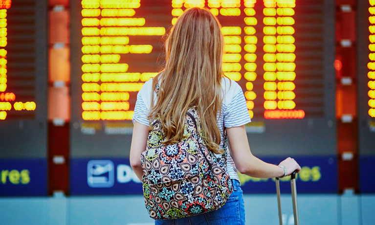 Back of a woman with a backpack and suitcase, looking at a flight information board in an airport.