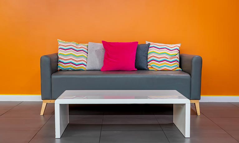 A grey sofa with colourful cushions against a bright orange painted wall.