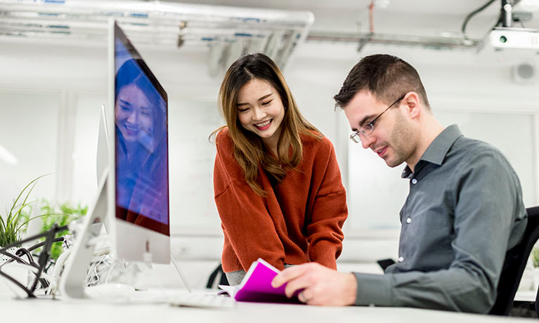 asian female student and male student looking at a text book in front of computer screen
