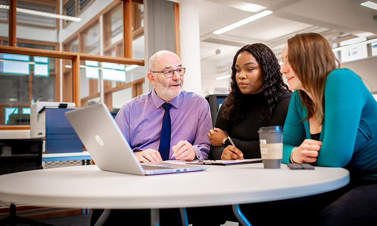 Two students sat talking to a lecturer with a laptop