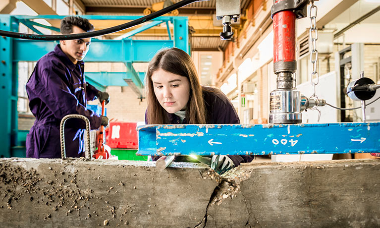 Female student working in structures lab with male student looking on.