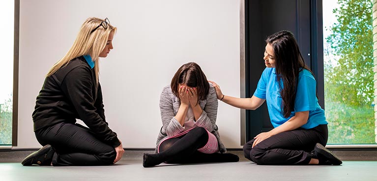 A patient sat on the floor with face in her palms and a nurse either side of her