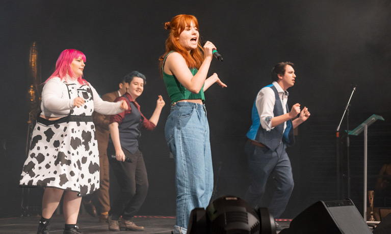 A student singing on a stage with backing singers and dancers.