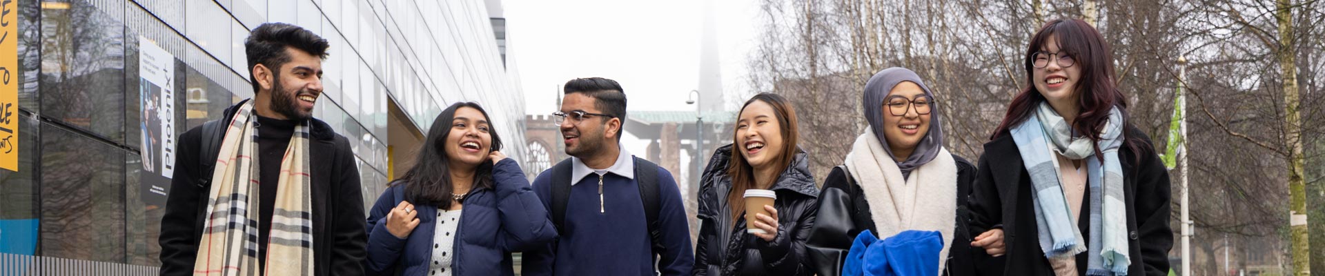 A group of students chatting and laughing as they walk past The Hub on campus with Coventry Cathedral behind them