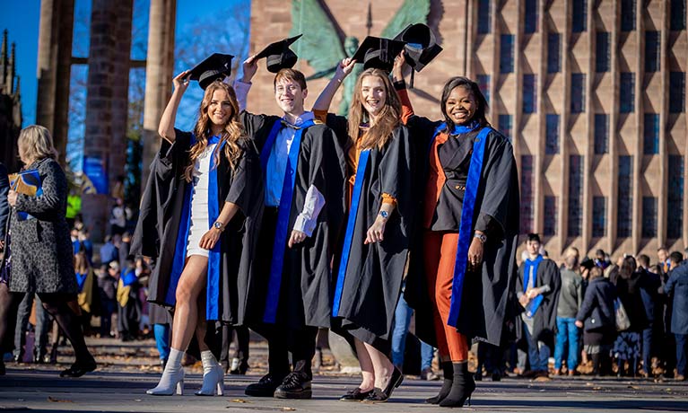 4 students smiling wearing graduation outfits standing outside Coventry cathedral